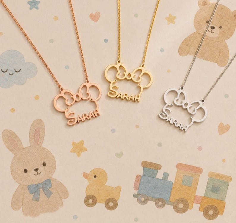 Op de afbeelding: Vier kettingen met de naam "SARAH" in een scriptlettertype. De hangers hebben de vorm van een strik met een muisoren-ontwerp. De kettingen zijn in ros&eacute;goud, goud en zilver. De achtergrond heeft een pastel babymotief met cartoon dieren.