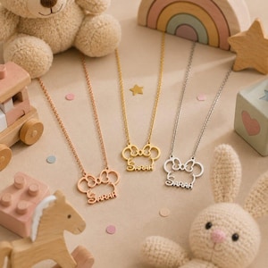 Pu&ograve; includere: Tre collane con un ciondolo a forma di topo e il nome "Sarah" in corsivo. Le collane sono in oro rosa, oro e argento. L'immagine include un orsacchiotto, giocattoli di legno e un coniglietto di peluche.