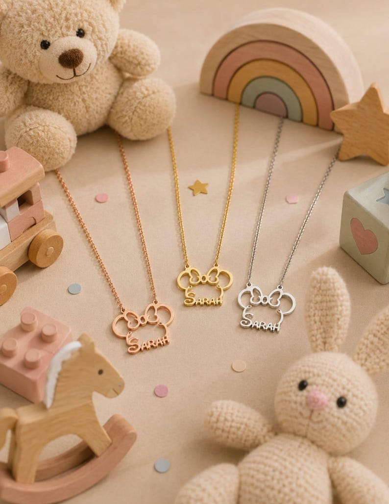 Pu&ograve; includere: Tre collane con un ciondolo a forma di topo e il nome "Sarah" in corsivo. Le collane sono in oro rosa, oro e argento. L'immagine include un orsacchiotto, giocattoli di legno e un coniglietto di peluche.