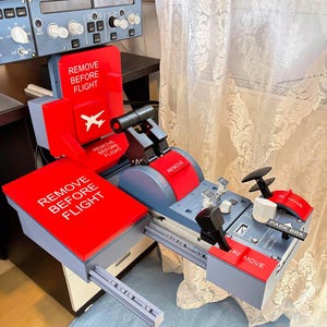 Op de afbeelding: Een vluchtsimulator throttle quadrant met grijze en rode onderdelen en meerdere rode labels met de tekst REMOVE BEFORE FLIGHT. De unit bevat zwarte gashendels, een parkeerremhendel en diverse bedieningsknoppen en schakelaars.