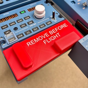Puede incluir: Un panel de control de radio de avión azul con una cubierta de plástico roja que tiene impreso el texto blanco REMOVE BEFORE FLIGHT. El panel incluye varios botones etiquetados como NAV, VOR, ILS, GLS, MLS, ADF, AM, HF y VHF, junto a un mando blanco.
