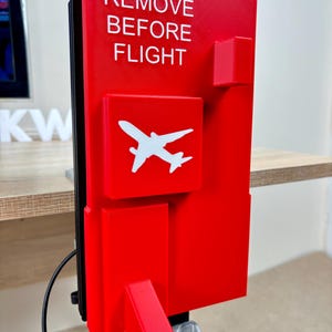 Op de afbeelding: Een rechthoekig rood 3D-geprint bord met de witte tekst REMOVE BEFORE FLIGHT en een wit vliegtuigicoon. Het bord heeft een verhoogd oppervlak met geometrische uitsteeksels en is gemonteerd op een metalen voet met een zwarte kabel aan de zijkant.