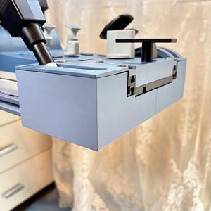 Könnte beinhalten: Eine hellblaue, rechteckige, 3D-gedruckte Kunststoff-Zubehörablage für eine Maschine. Sie verfügt über einen schwarzen Düsenaufsatz, eine weiße zylindrische Komponente und einen schwarzen T-förmigen Griff auf einer flachen, modularen Oberfläche.