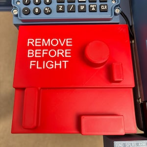 May include: A bright red rectangular cover featuring the white text REMOVE BEFORE FLIGHT, positioned below a blue control panel with a numeric and letter keypad. The cover includes a circular knob and two rectangular protrusions on its surface.