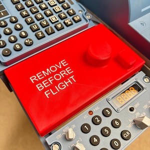 Op de afbeelding: Een felrode rechthoekige afdekplaat met de witte tekst REMOVE BEFORE FLIGHT, geplaatst over een lichtblauw bedieningspaneel van een vliegtuig met een numeriek toetsenbord, draaischakelaars en een klein digitaal display met vier oranje cijfers.