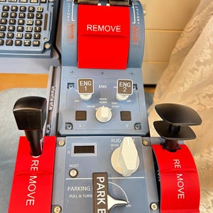 Puede incluir: Un cuadrante de aceleración para simulador de vuelo con un panel de control gris, interruptores ENG 1 y ENG 2, una palanca de freno de estacionamiento etiquetada PARK BRK y tres palancas rojas con el texto REMOVE impreso en blanco.