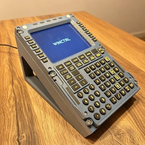 Op de afbeelding: Een grijze replica van een vluchtmanagementcomputer met een blauw scherm met de tekst WINCTRL, een volledig QWERTY-toetsenbord met ronde knoppen en extra functietoetsen aan de zijkanten, gemonteerd op een schuine steunvoet.