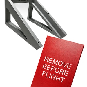 Puede incluir: Un soporte de estante triangular de metal gris con el texto MrKwork impreso en el lateral, acompañado de un letrero rectangular rojo con el texto blanco REMOVE BEFORE FLIGHT. El soporte tiene orificios pretaladrados para el montaje.
