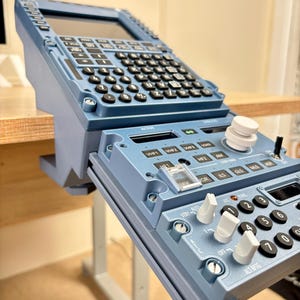 Puede incluir: Una unidad de control de sistema de gestión de vuelo de avión en color azul que cuenta con una pantalla superior, un teclado alfanumérico completo y un panel inferior con varios mandos, interruptores y botones con etiquetas de aviación.