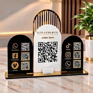Può includere: Un espositore in acrilico color oro, nero e bianco con tre pannelli ad arco. Il pannello centrale bianco mostra "ELITE GEMS & CO. order here" e un grande codice QR. I due pannelli laterali neri presentano icone dorate e codici QR per i social.