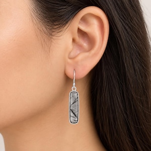 May include: A silver drop earring featuring a rectangular, translucent stone with visible black needle-like inclusions. The stone is set in a smooth silver frame and hangs from a simple silver hook wire, creating a clean, elongated design.