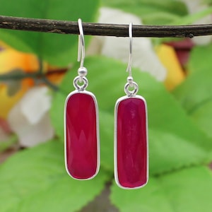 May include: A pair of magenta-coloured rectangular drop earrings with silver-coloured metal frames. Each earring has a simple hook and a small decorative ball. The background is blurred with green and yellow hues.