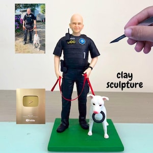以下が含まれることがあります： 黒いリングを持った警察官と白い犬の粘土彫刻。 警官は黒い制服を着て、赤いリードを持っています。 彫刻は緑色の台座にあります。 金色のプラークと写真が背景にあります。