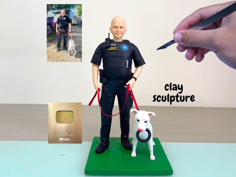 以下が含まれることがあります： 黒いリングを持った警察官と白い犬の粘土彫刻。 警官は黒い制服を着て、赤いリードを持っています。 彫刻は緑色の台座にあります。 金色のプラークと写真が背景にあります。