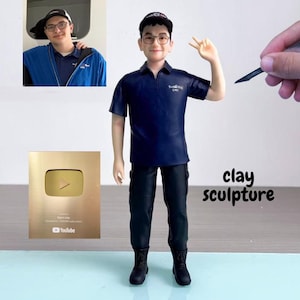 Könnte beinhalten: Ton-Skulptur einer Person in einem marineblauen Poloshirt, schwarzer Hose und Stiefeln. Die Skulptur hält zwei Finger hoch. Ein goldener YouTube-Play-Button und ein Foto befinden sich im Hintergrund. Die Worte "clay sculpture" sind auf dem Bild geschrieben.