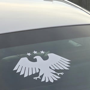 May include: White vinyl decal of an eagle with outstretched wings and three stars above its head, affixed to the rear window of a vehicle. The eagle design is a silhouette, creating a bold, graphic statement.