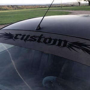 Puede incluir: Coche negro con una pancarta personalizada en el parabrisas. La pancarta presenta la palabra "custom" en una fuente estilizada con elementos decorativos. La antena del coche es visible, y el fondo muestra un campo verde y árboles.