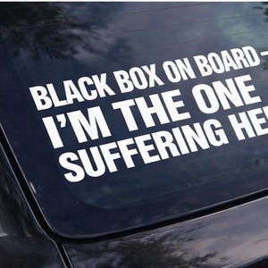 May include: White vinyl decal applied to a car window. The text reads "BLACK BOX ON BOARD - I'M THE ONE SUFFERING HERE" in a bold, sans-serif font. The decal is likely for humor or self-identification.