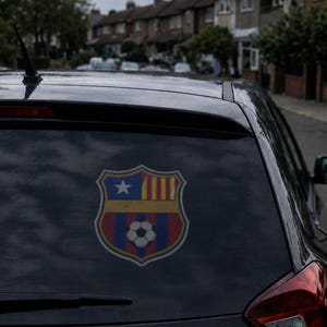 May include: A dark gray car rear window featuring a decal of a shield-shaped emblem. The emblem displays a blue section with a white star, a yellow section with four vertical red stripes, and a lower section with blue and red stripes and a soccer ball.