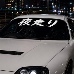 Op de afbeelding: Een witte sportwagen met een grote witte sticker op de voorruit met de Japanse kanji-tekens 夜走り in een penseelstreekstijl. Het voertuig heeft een geventileerde motorkap, zwarte zijspiegels en een achterspoiler, gefotografeerd in de nacht.