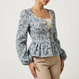 May include: A light blue floral patterned jacket with a square neckline and long sleeves. The jacket has a peplum waist and is buttoned up the front. The model is wearing a white tank top and tan pants.
