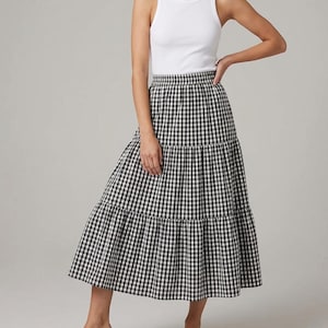 May include: A white ribbed tank top paired with a black and white checkered tiered midi skirt. The skirt features a classic gingham pattern and a flowing silhouette, creating a casual yet stylish ensemble. The outfit is suitable for warm weather.