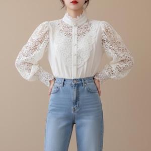 Puede incluir: Una blusa blanca con cuello alto con encaje y delicados detalles de encaje en la parte delantera y las mangas. La blusa presenta botones perlados y se combina con vaqueros de mezclilla azul claro. Las mangas son largas y abullonadas.