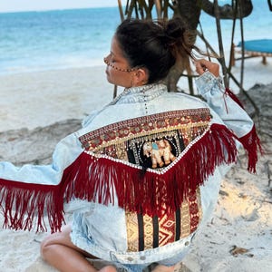May include: Light blue denim jacket with detailed embellishments. The back has a decorative panel with an elephant motif, gold accents, and red fringe. The jacket is adorned with various patterns and textures, creating a bohemian aesthetic.