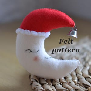 May include: A white felt crescent moon with closed eyes and a small pink cheek. It wears a red hat with white trim and a silver bell. The text "Felt pattern" is printed on the image.