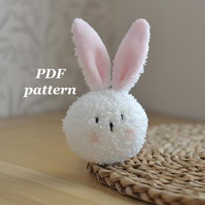 May include: A white, fluffy bunny ornament with pink inner ears and rosy cheeks. The bunny is made of a soft, textured material and has small black eyes. The text "PDF pattern" is visible in the image. The bunny is sitting on a woven mat.