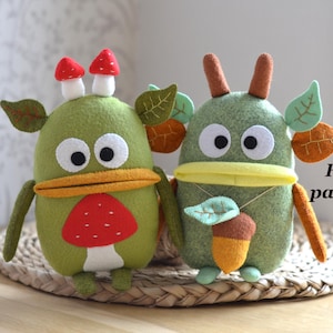 May include: Two whimsical felt creatures in shades of green, adorned with leaf and mushroom details. One has a red mushroom on its head and a mushroom on its belly. The other has an acorn pendant. The text "Felt pattern" is visible.