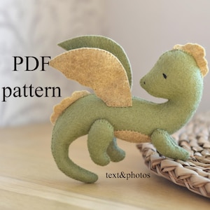 以下が含まれることがあります： 緑とベージュの色合いの手作りフェルトドラゴン。ドラゴンは翼ととげのあるとさかがあり、織られた表面に置かれています。画像には「PDF pattern」と「text&photos」の文字が含まれています。