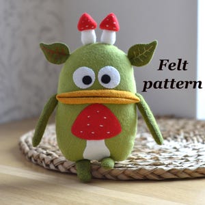 May include: A handmade green felt monster with white eyes, a yellow beak, and a red mushroom design on its chest. Two red and white mushroom-shaped accents sit atop its head. The text "Felt pattern" is visible.