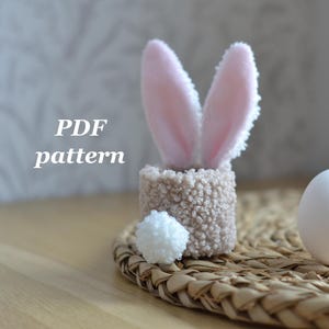 Peut inclure: Un projet artisanal sur le thème du lapin de Pâques. Il comprend un petit récipient texturé de couleur beige avec des oreilles de lapin roses et blanches, une queue en pompon blanc et un œuf blanc. Le texte "PDF pattern" est visible.