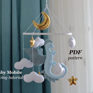 May include: A baby mobile featuring a light blue dragon, gold stars, a gold crescent moon, and white clouds. The mobile is suspended from a white circular frame. Text on the image reads "Baby Mobile sewing tutorial" and "PDF pattern".
