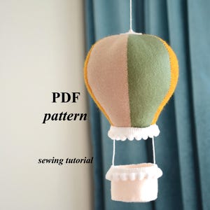 Puede incluir: Móvil de globo aerostático hecho a mano con fieltro. El globo está dividido en secciones beige, verdes y amarillas. La cesta es de color rosa claro con ribete blanco. El texto "PDF pattern sewing tutorial" es visible.