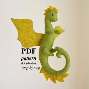 May include: A handmade felt dragon in shades of green and yellow. The dragon has large yellow wings, a spiky yellow crest, and a curled tail. The text "PDF pattern 45 photos step by step" is displayed below the dragon.