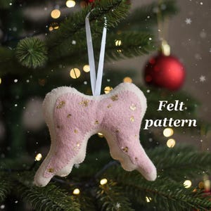 May include: A pink felt angel wing ornament with gold glitter accents hangs from a white ribbon on a Christmas tree. The text "Felt pattern" is visible. Red ornaments and twinkling lights are in the background.