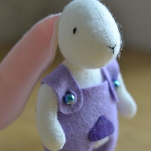 May include: A handmade white bunny plush toy wearing a purple felt jumpsuit with iridescent blue buttons. The bunny has a pink inner ear and a stitched black nose and mouth. A small, purple felt pocket is sewn on the front of the jumpsuit.