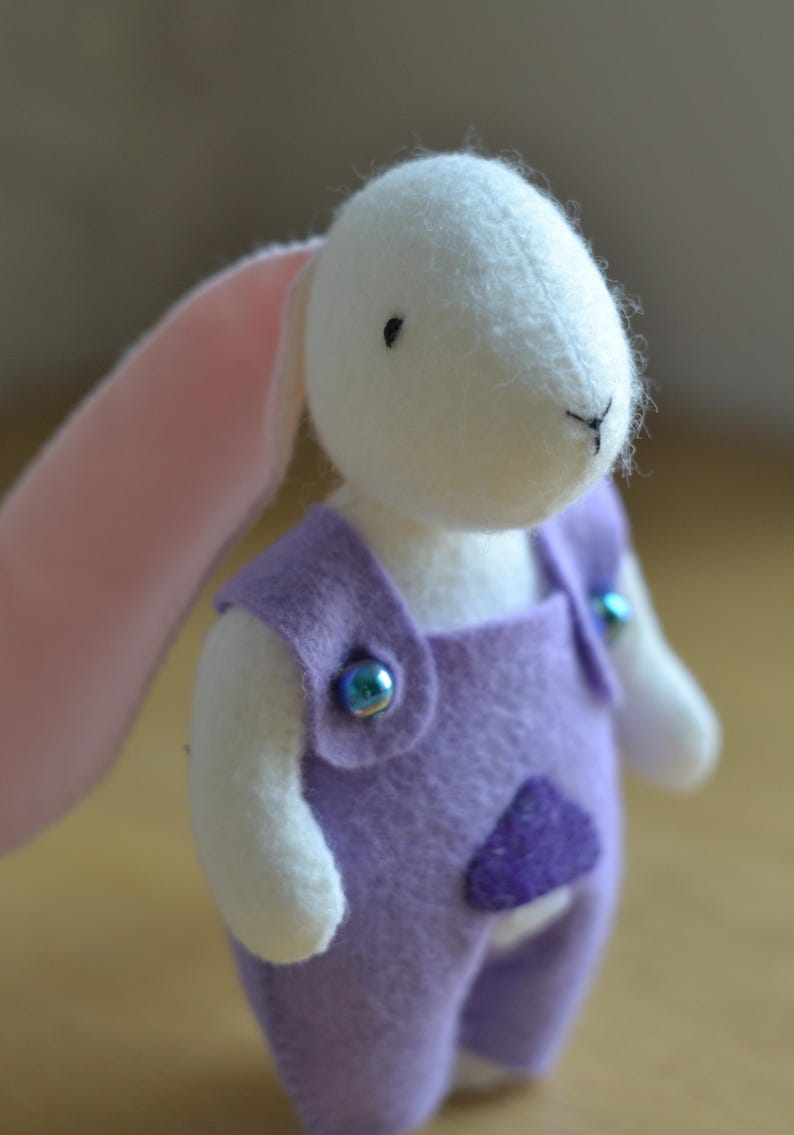 May include: A handmade white bunny plush toy wearing a purple felt jumpsuit with iridescent blue buttons. The bunny has a pink inner ear and a stitched black nose and mouth. A small, purple felt pocket is sewn on the front of the jumpsuit.