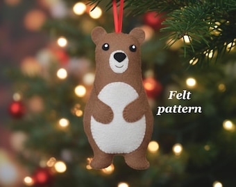 May include: A brown felt bear ornament with a white belly, black eyes, and a smiling mouth. The ornament hangs from a red ribbon against a blurred Christmas tree background. Text on the right says "Felt pattern".