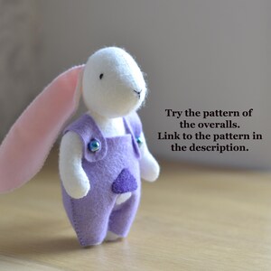 May include: A handmade plush bunny with a white head, pink ears, and a lavender overall. The overall has a purple heart-shaped patch and is fastened with blue buttons. The text on the image says, "Try the pattern of the overalls. Link to the pattern in the description."