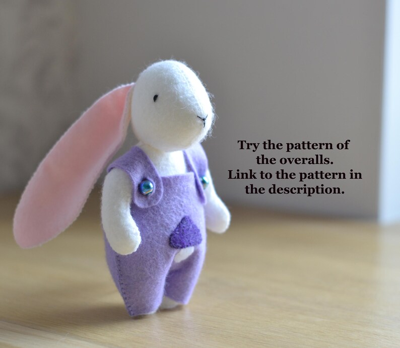 May include: A handmade plush bunny with a white head, pink ears, and a lavender overall. The overall has a purple heart-shaped patch and is fastened with blue buttons. The text on the image says, "Try the pattern of the overalls. Link to the pattern in the description."