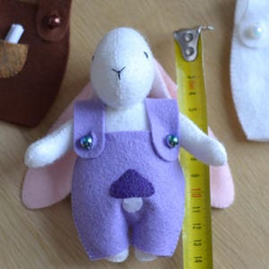 May include: A handmade felt bunny doll wearing purple overalls with a mushroom applique. The bunny is white with pink ears and is approximately 4 inches tall. The overalls have decorative buttons. A measuring tape is next to the doll.