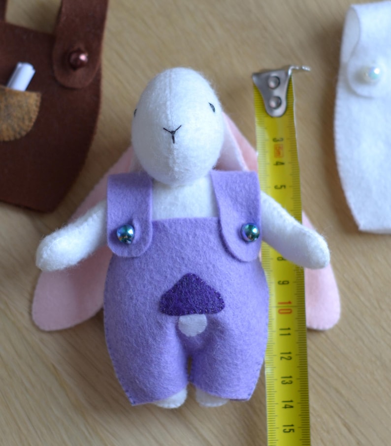 May include: A handmade felt bunny doll wearing purple overalls with a mushroom applique. The bunny is white with pink ears and is approximately 4 inches tall. The overalls have decorative buttons. A measuring tape is next to the doll.