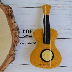 May include: A felt ukulele in shades of yellow, with black and white details. The ukulele has a circular sound hole and four white strings. Text on the image reads "PDF pattern sewing tutorial".