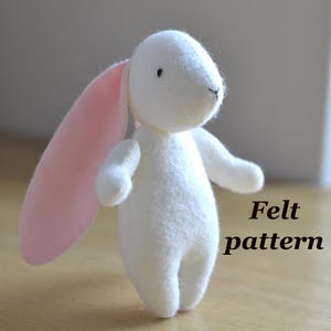 May include: A white felt bunny with pink ears and small black eyes. The bunny has outstretched arms. The text "Felt pattern" is on the right. The bunny is on a wooden surface.