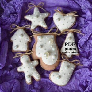Pode incluir: Uma coleção de enfeites de Natal de feltro feitos à mão em várias formas, incluindo uma estrela, um coração, uma casa, uma luva, um boneco de gengibre, uma árvore e uma meia. Cada enfeite é branco com enfeites de pérolas e um laço de barbante para pendurar. Os enfeites são exibidos em uma caixa com papel de seda roxo.