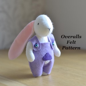 May include: A handmade felt bunny doll wearing purple overalls with blue button accents. The bunny has long pink ears and white arms and legs. The text "Overalls Felt Pattern" is displayed in the image.