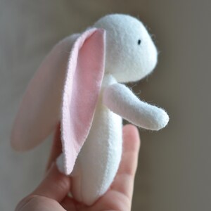 May include: A handmade white stuffed bunny with pink inner ears. The plush toy has a small black eye and is held in a hand. The bunny is made of soft fabric and is a cute, whimsical design.